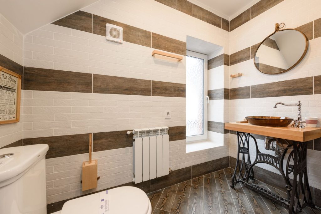 Bathroom with a small exhaust fan on the upper wall, a window, a radiator and a round mirror