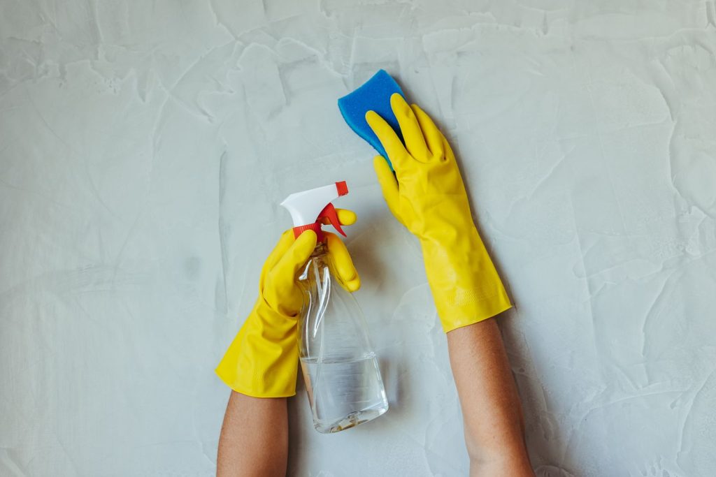 Hands in yellow rubber gloves holding a sponge and a spray bottle against a pale plastered wall