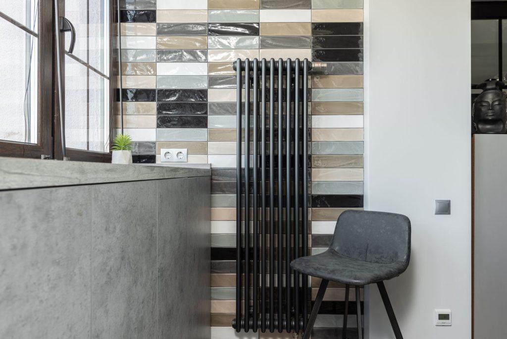 Zwarte verticale radiator naast een raam tegen een muur met kleurige tegels en een grijze stoel