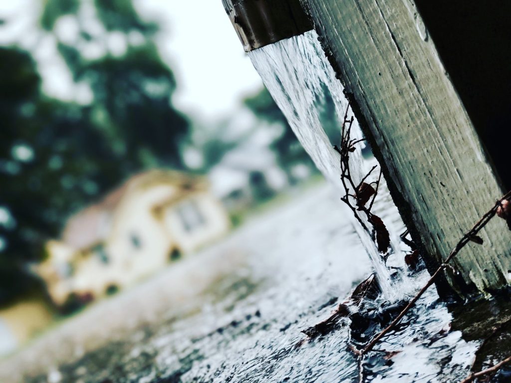 Rainwater pouring from the end of a roof gutter onto the ground next to a house
