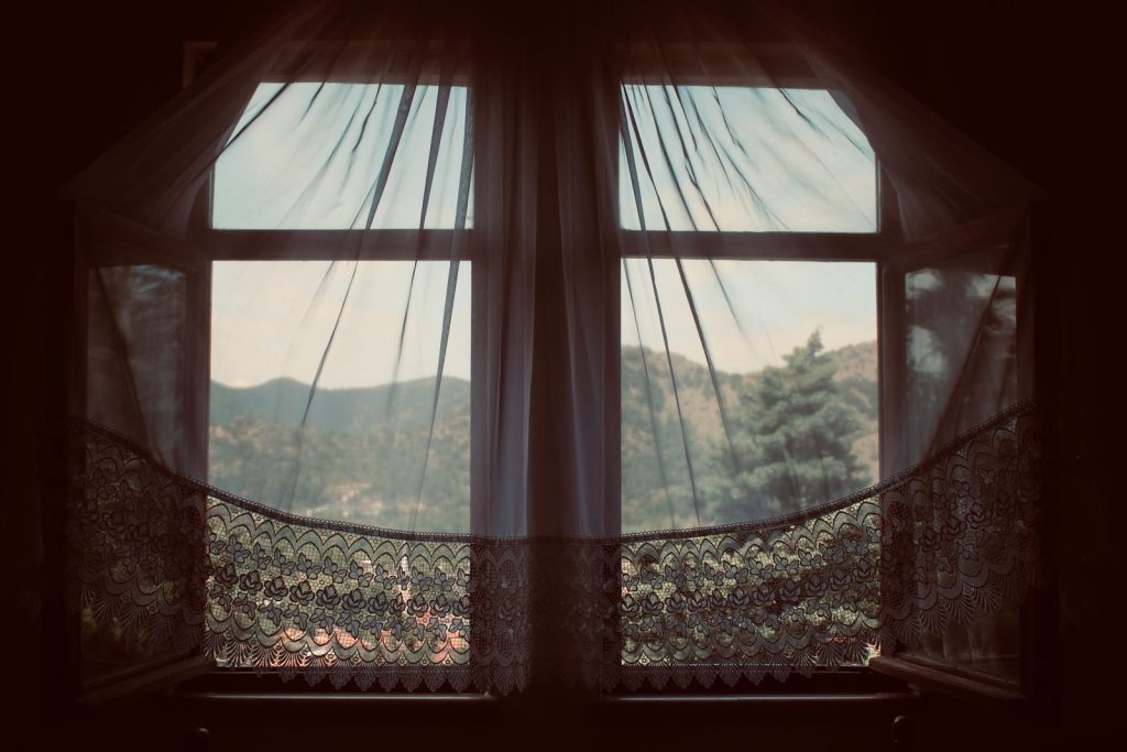 Open double window with sheer lace curtains and a view of green hills outside