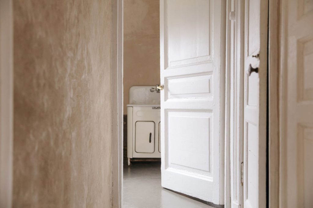 Doorkijk door een half openstaande witte deur naar een oude witte wastafel in een eenvoudig interieur