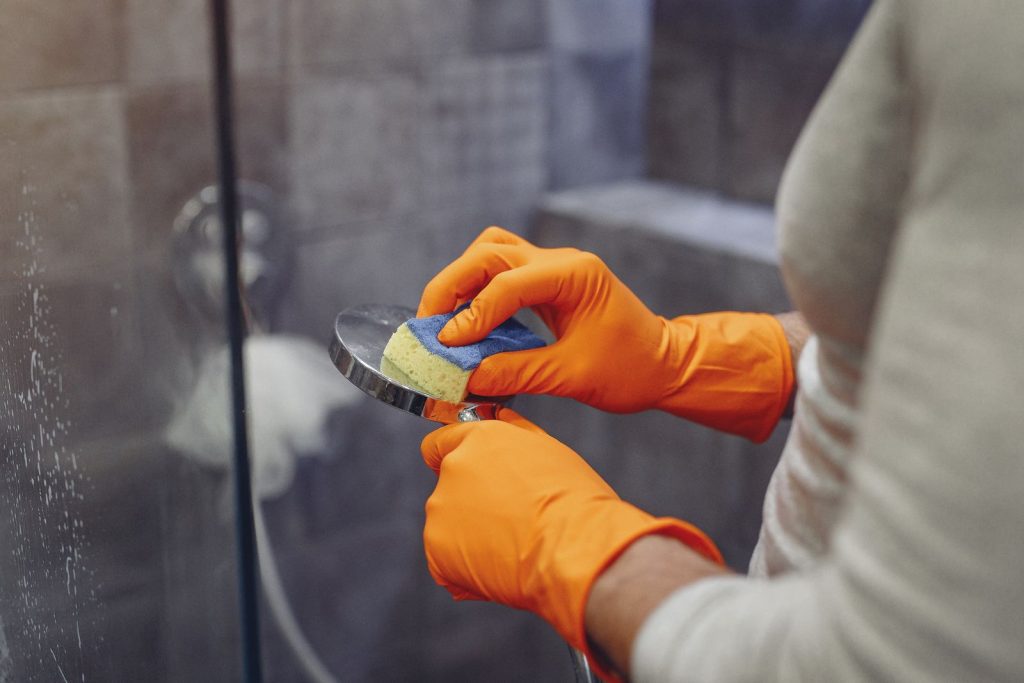Hands in orange rubber gloves cleaning a chrome shower head with a sponge