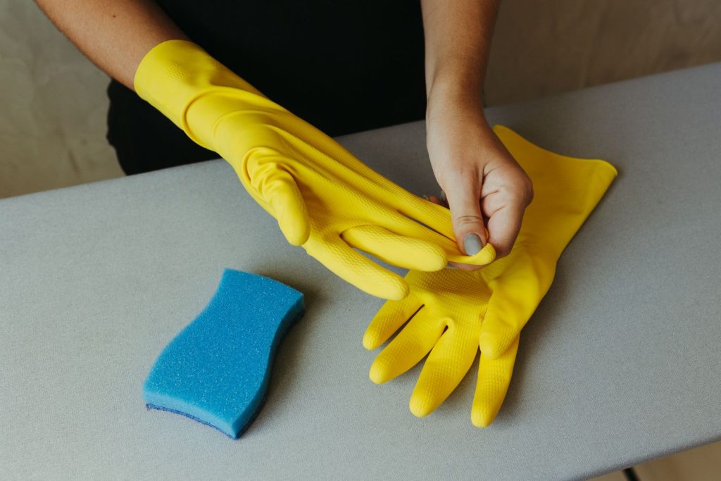 Handen trekken gele rubberen schoonmaakhandschoenen aan naast een blauwe spons op een grijze tafel