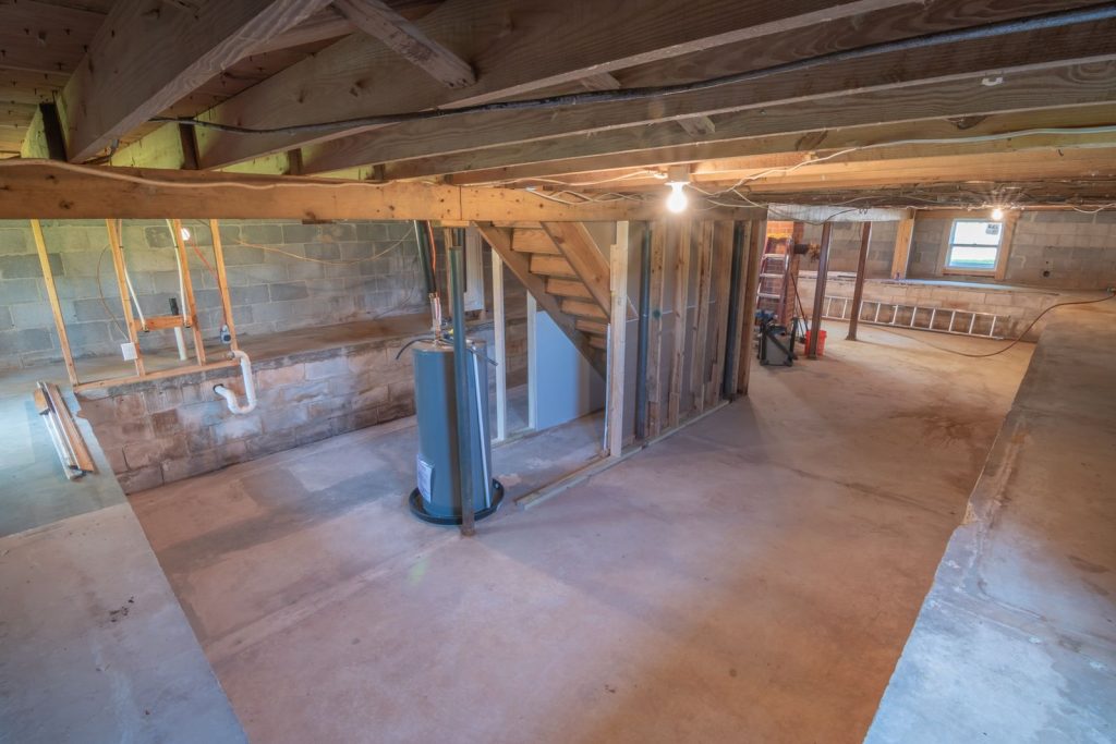 Unfinished basement with concrete block walls, exposed pipes, a water heater and wooden stairs